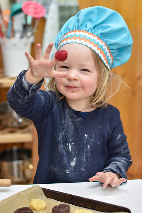 Ein Kind mit Kochmütze steht beim Backen und lächelt fröhlich in die Kamera.
